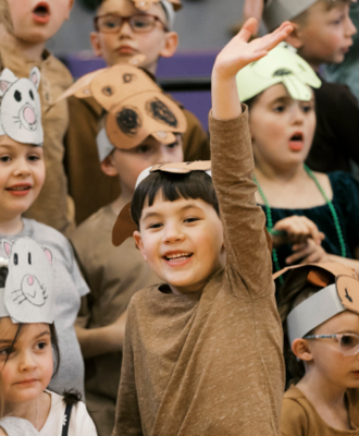 A close-up of a group of children, in the foreground a young boy smiles broadly with his arm raised.
